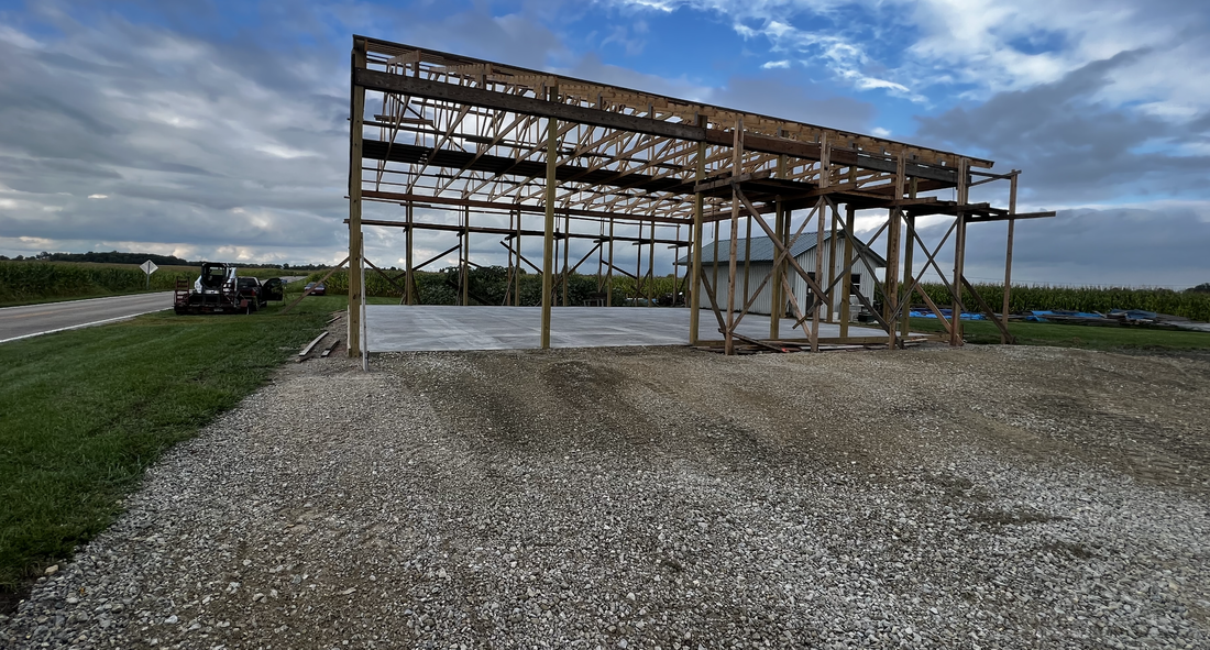 A construction site featuring a large concrete pad with a smooth finish, intended for a metal framework structure overhead, possibly a future building or barn. There is construction equipment on the left and a gravel path leading to the site. The background shows a rural setting with open fields, a road, and a cloudy sky.