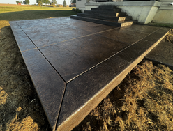 An angled view of a chocolate-brown stamped concrete patio that emulates large slate tiles, extending towards a set of stairs. The area surrounding the patio is covered with straw, and a residential lawn can be seen in the background under a clear sky.