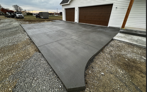 A newly constructed concrete driveway in a residential area with a smooth, dark gray finish. The driveway is divided into large rectangular sections by control joints and is flanked by a two-car garage with white walls and brown doors on one side, and a gravel area transitioning to bare soil on the other. In the background, overcast skies loom above, with construction trailers and vehicles parked on the adjacent gravel lot.